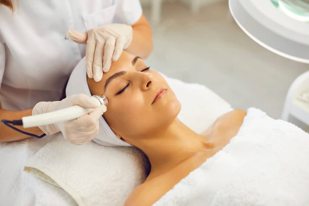 Esthéticienne en gants appliquant un traitement de radiofréquence ou hydrafacial sur le visage d'une cliente détendue enroulée dans une serviette blanche.