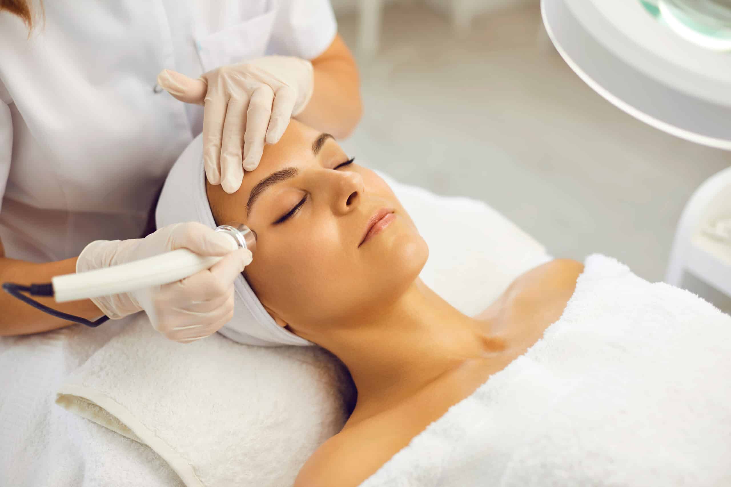 Esthéticienne en gants appliquant un traitement de radiofréquence ou hydrafacial sur le visage d'une cliente détendue enroulée dans une serviette blanche.