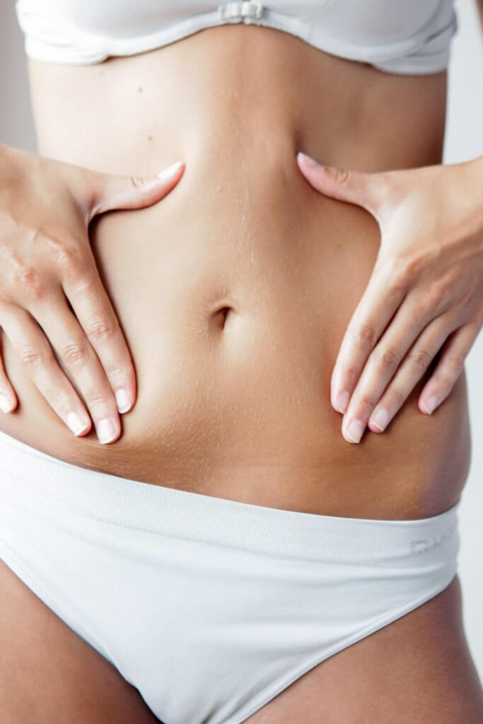 Gros plan sur un ventre féminin tonique, mains tenant doucement les flancs, montrant un nombril et un duvet fin, vêtue d'un sous-vêtement blanc.